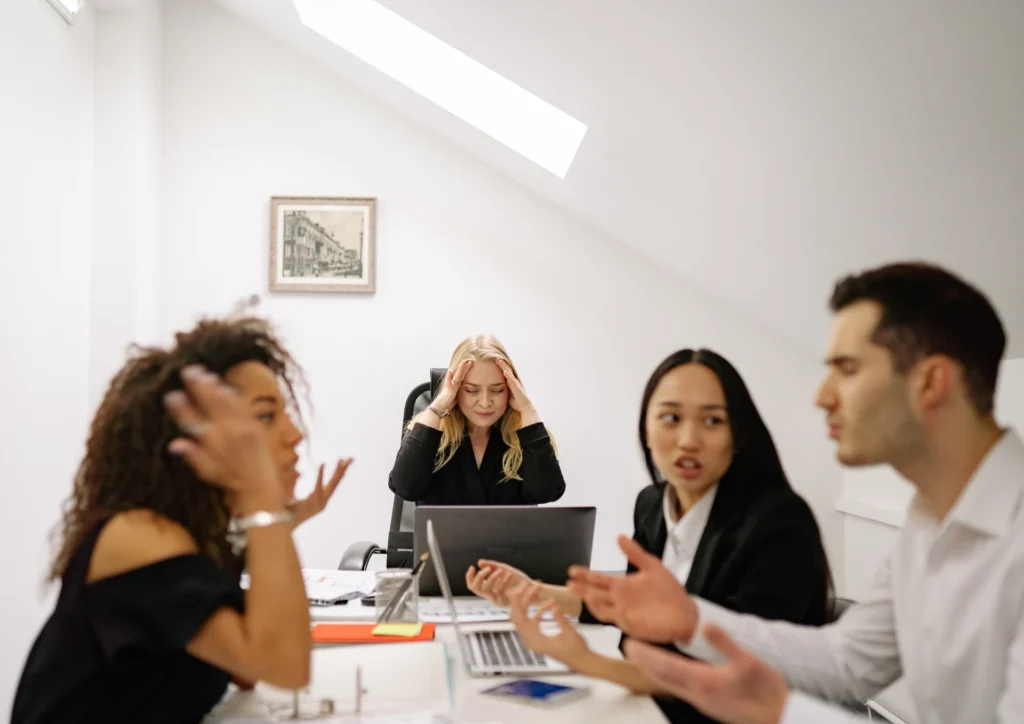 In a bright office, a stressed woman sits at a laptop, holding her head. Three colleagues engage in a heated discussion, conveying tension and disagreement