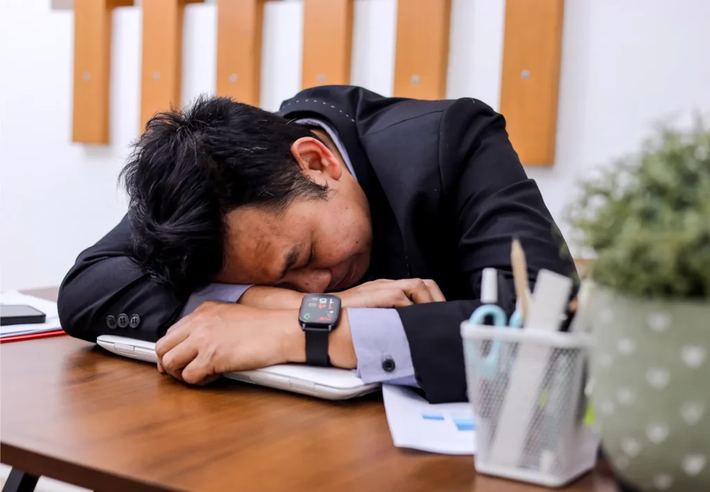 Man in a suit asleep on a laptop at a wooden desk, wearing a smartwatch. Office supplies and a plant are visible, conveying fatigue and stress.