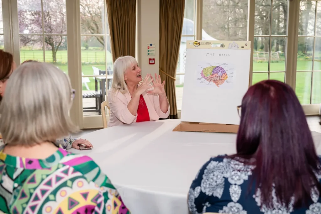 Nicola Hunt engages a small group in a discussion about the brain using a colorful diagram on an easel in a bright room with large windows and garden views