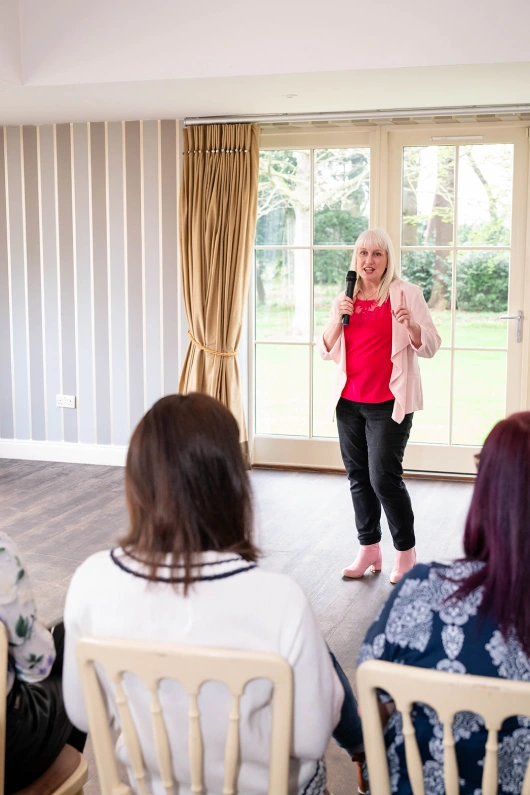 Nicola Hunt with a microphone speaks confidently to an audience in a bright room. She wears a pink blouse and blazer, creating an engaging atmosphere